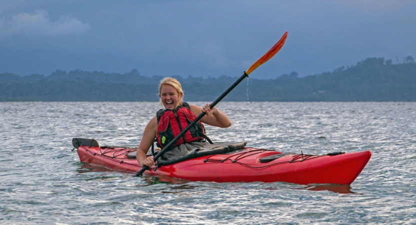 A person paddling a red kayak on open water smiles. 